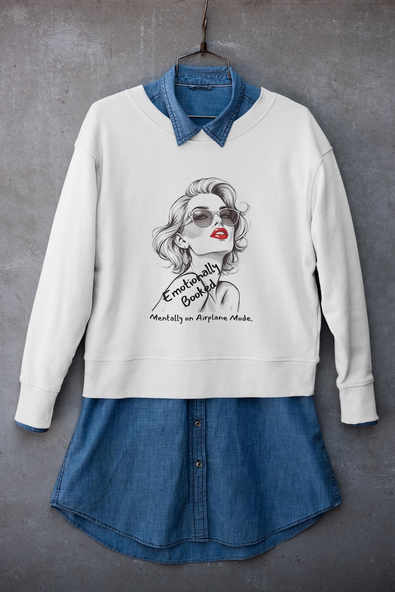 May include: White sweatshirt with a graphic of a woman wearing sunglasses and red lipstick. The text reads "Emotionally Booked, Mentally on Airplane Mode." The sweatshirt is layered over a denim shirt.