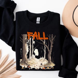 Könnte beinhalten: Schwarzes Sweatshirt mit einem orangefarbenen "FALL"-Motiv. Die Grafik zeigt eine Waldszene mit Bäumen und Herbstlaub. Ein beigefarbener Fedora-Hut ist im Hintergrund zu sehen. Das Sweatshirt liegt auf einer weißen Oberfläche.