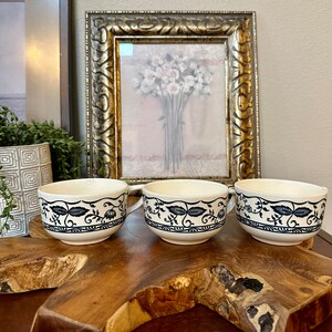 May include: Three white ceramic teacups with black floral designs and gold trim. The cups are sitting on a wooden surface.