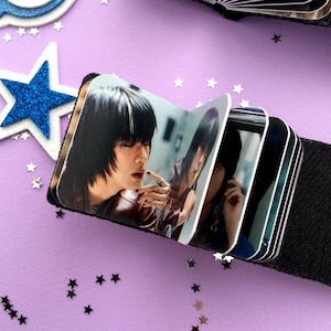 May include: A small, black photo album with a series of images of a person with black hair. The album is open, revealing several pages with the photographs. The background is a light purple color with star-shaped confetti.