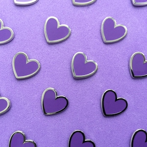 May include: A collection of purple heart-shaped enamel pins with silver edging, arranged on a textured, light purple surface. The pins have a shiny finish. The background is a plain, light purple colour.