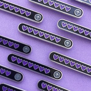 Peut inclure: Épingles en émail avec un fond noir, une bordure argentée et des icônes de cœur violet. Chaque épingle comporte une icône de flèche à une extrémité. Les épingles sont disposées sur une surface violette texturée.