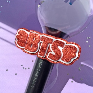 Pode incluir: Um bastão de luz preto com um topo esférico transparente e um acessório com o logótipo BTS em glitter vermelho. O cabo apresenta o texto OF THE SOUL em letras brancas. O design inclui um contorno branco à volta das letras com glitter.