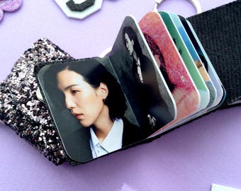 May include: A small, black fabric photo album with a silver keyring. The album contains several color photographs. The album is resting on a purple surface, with decorative glittery accents and felt letters in the background.