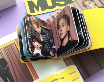May include: A small, open photo album featuring images of a musician. The album is surrounded by a yellow fabric and a yellow and black foam cutout that reads "JIMIN MUSE". The album is open, displaying several photos.