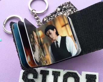 May include: A small photo album keychain featuring images of a person. The album is attached to a silver keyring and has a black fabric cover. The word "SUGA" is spelled out in black and white glitter letters.