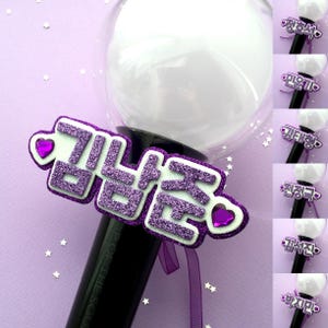 May include: A purple glittery name tag with Korean text is attached to a black light stick. The tag has a white border and heart-shaped accents. Additional light sticks with different names are visible.