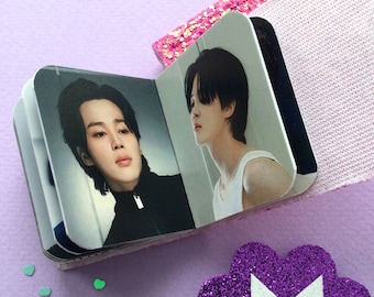 May include: A small, open photo album featuring images of a person with dark hair. The album is on a light purple surface, next to a pink glittery accessory and a purple glittery logo.