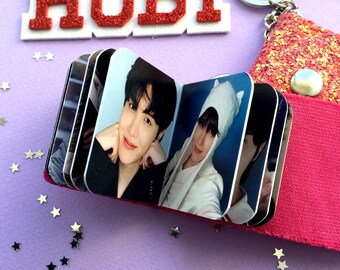 May include: A small photo album with images of a person, held together by a pink fabric. The album is next to a keychain with a glittery pink charm and a red glitter sign that says "HOBI". The background is purple.