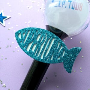 May include: A teal glittery fish-shaped accessory with the word "SUPERKIND" cut out, attached to a black handle. The top of the handle has a clear, spherical object with the text "LP. TOUR". The background is a light purple with star-shaped confetti.