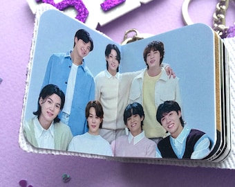 May include: A small, open photo album featuring a group of seven people. The album is white with a silver-colored chain and a purple glittery charm that reads "BTS". The photo shows the group in various pastel-colored outfits.