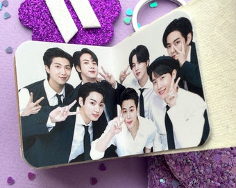 May include: A small, open booklet featuring a photograph of seven individuals in formal attire, including white shirts and black ties, posing with peace signs. The booklet has a purple glitter cover and is accented by a white BTS logo emblem.