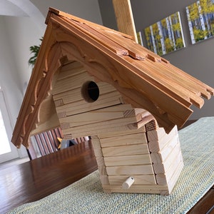 Può includere: Una casetta per uccelli in legno con un tetto a tegole e una piccola apertura per consentire agli uccelli di entrare. La casetta è realizzata in legno marrone chiaro e presenta un design decorativo sul tetto.