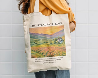 Könnte beinhalten: Eine weiße Canvas-Tragetasche mit einem Landschaftsgemälde eines Blumenfeldes und einem Haus in der Ferne. Der Text "THE STEADFAST LOVE" ist oben auf der Tasche aufgedruckt. Das Zitat "of the Lord never cease, his mercies never come to an end; they are new every morning; great is your faithfulness." ist unter dem Bild aufgedruckt. Der Text "LAMENTATIONS 3:22-23" ist unter dem Zitat aufgedruckt.