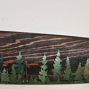 May include: A wooden oar painted with a forest scene. The oar has a dark brown stain and is decorated with green trees. The oar has several hooks on the side.