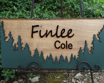 May include: A wooden sign with a green forest silhouette at the bottom. The sign has the names "Finlee Cole" written in black cursive lettering.