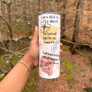May include: A white tumbler with floral and abstract designs, featuring the text "I am a child of God" and Bible verses. The tumbler has a silver lid and is held against a blurred outdoor background.