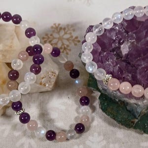 Puede incluir: Dos pulseras hechas con cuentas de amatista, piedra lunar y cuarzo rosa. Las pulseras se muestran sobre un lecho de cristales blancos y morados.