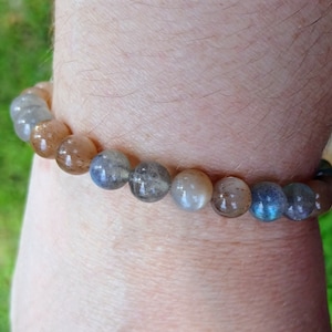 Puede incluir: Un brazalete de cuentas redondas e iridiscentes en tonos grises, marrones y azules. El brazalete se lleva en la muñeca, mostrando las variaciones naturales y los colores sutiles de las piedras. Las cuentas miden aproximadamente 0,8 cm de diámetro.