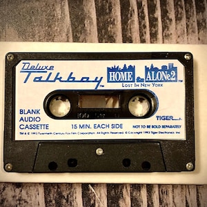 Puede incluir: Un casete de audio en blanco y negro con la etiqueta "Deluxe Talkboy" y la marca "Home Alone 2". El casete tiene el texto "Blank Audio Cassette" y "15 min. por lado". Sobre un fondo blanco.