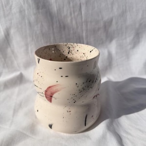 Puede incluir: Un jarrón de cerámica color crema con una forma redondeada única. El jarrón está decorado con salpicaduras y pinceladas de pintura negra y roja, añadiendo un toque artístico. El interior también es color crema.