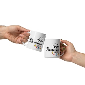 May include: Two white ceramic mugs with the text "The Grandfather" and a black illustration of marionette puppets. The mugs also feature colorful toy accents. The mugs are held up in a celebratory gesture.