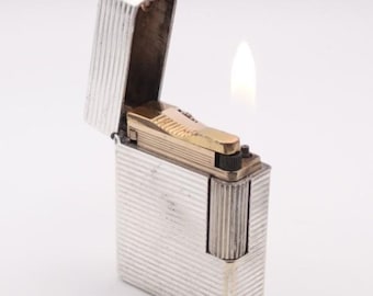 Könnte beinhalten: Ein silbernes Feuerzeug mit geripptem Design und goldfarbenem Oberteil. Das Feuerzeug ist geöffnet, mit einer sichtbaren Flamme. Auf der Oberseite des Feuerzeugs ist der Buchstabe "M" eingraviert.