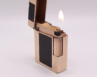 Könnte beinhalten: Ein goldfarbenes Feuerzeug mit rechteckiger Form und einer schwarzen Platte auf der Vorderseite. Das Feuerzeug ist geöffnet, mit einer sichtbaren Flamme. Die Oberseite des Feuerzeugs hat ein kleines Emblem. Das Feuerzeug hat eine gerippte Textur.