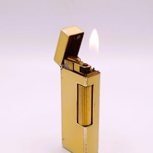 May include: A gold-coloured lighter with a rectangular body and a hinged top. The lighter is open, and a flame is visible. The lighter has a textured, ribbed design on the front. The background is a solid white.