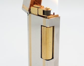 Könnte beinhalten: Ein silberner und goldener Feuerzeug mit Flamme. Das Feuerzeug hat eine rechteckige Form mit vertikalen Goldstreifen und einem goldenen Akzent auf der Vorderseite. Die Oberseite ist offen und gibt den Blick auf den Innenmechanismus frei.