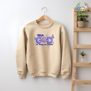 Pu&ograve; includere: Una felpa beige con un design floreale che dice "With God all things are possible".