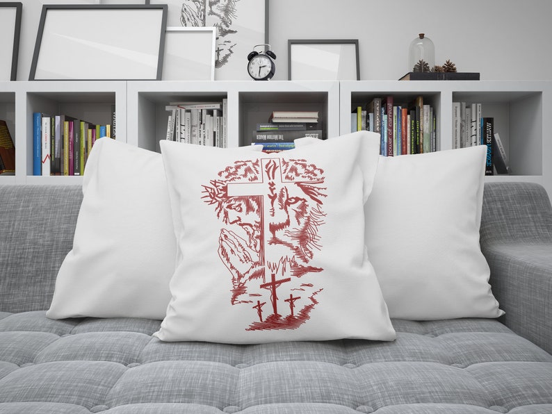 K&ouml;nnte beinhalten: Ein wei&szlig;es Kissen mit einem roten gestickten Design aus einem Kreuz, einem L&ouml;wenkopf und einer betenden Figur. Das Design ist von einer Dornenkrone umgeben.