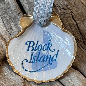 May include: A white seashell ornament with a gold-painted edge and a blue and white design. The ornament has the words "Block Island" in blue script. A gray and pink ribbon is attached for hanging.