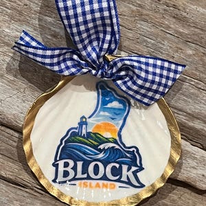 May include: A decorative seashell ornament featuring a Block Island design. The white shell has a gold-painted edge and a blue and white gingham ribbon. The design includes a lighthouse, waves, and the text "BLOCK ISLAND".