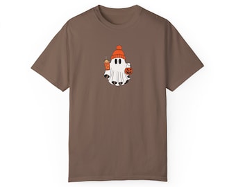 May include: A brown short-sleeve t-shirt featuring a central graphic of a white ghost wearing an orange beanie. The ghost holds an orange pumpkin bucket in one hand and a tall orange beverage cup with a straw in the other hand.