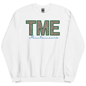May include: White sweatshirt with the letters "TME" in a leopard print design outlined in green. Below the letters, the word "Mountaineers" is written in blue script. The sweatshirt has a crew neck and long sleeves.