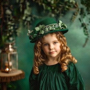 May include: A young girl with curly blonde hair wears a green hat with a floral embellishment and a matching green dress. The background is a green and brown backdrop with a lantern.