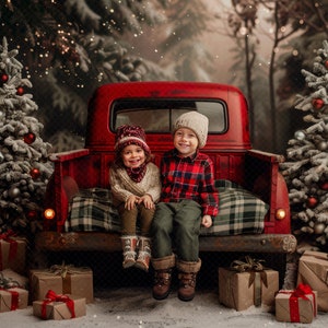 May include: Two children wearing winter hats and clothes sit in the back of a red pickup truck. The truck is decorated with Christmas lights and there are two snow-covered Christmas trees on either side of the truck. There are wrapped presents on the ground in front of the truck.