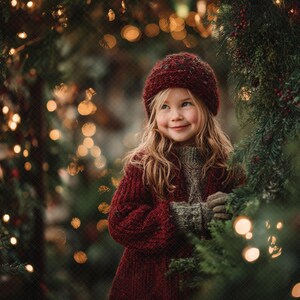May include: A young person wearing a burgundy knit hat, sweater, and coat, smiling amidst a festive holiday scene. The background features a garland of evergreen branches, twinkling lights, and bokeh effects, creating a warm, inviting atmosphere.