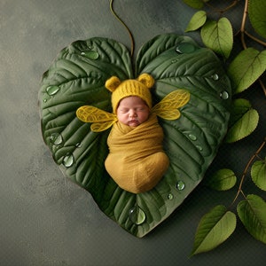 May include: A newborn baby wrapped in a yellow blanket and wearing a yellow bee-themed hat with wings, resting on a large green leaf with water droplets. The background is a muted gray-green with additional green leaves.