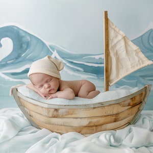 Pode incluir: Um recém-nascido usando um chapéu cor de creme dorme pacificamente em um barco de madeira. O barco é cercado por um tecido azul claro e um fundo pintado de ondas azuis.