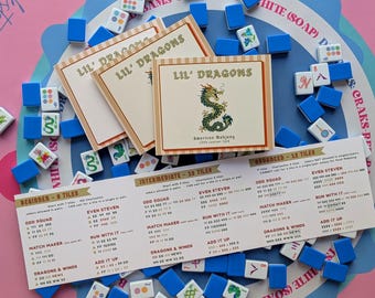 May include: A set of Lil' Dragons American Mahjong learner cards and a long instructional guide featuring game rules and tile combinations. The cards display a green dragon illustration and text, surrounded by scattered blue and white game tiles.