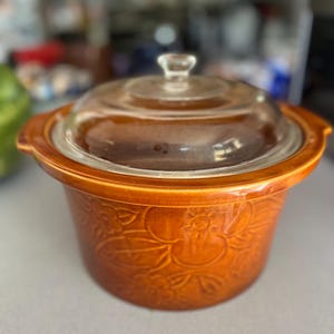May include: A vintage brown ceramic slow cooker with a clear glass lid. The slow cooker features a floral embossed design and two side handles. The lid has a small handle on top.