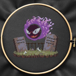 May include: A cross-stitch embroidery of a purple and black ghost Pokemon character, Gengar, floating above two gray tombstones with white bones on the ground.