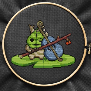 May include: Cross-stitch embroidery of a green and brown creature holding a blue and brown instrument, sitting on a green leaf with red berries.