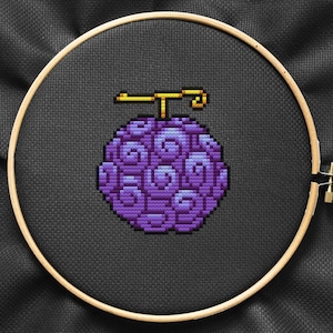 May include: A cross-stitch design of a purple, pixelated fruit, possibly a Devil Fruit, set within a wooden embroidery hoop. The fruit has a unique, swirling pattern and a golden stem. The background is a dark, textured fabric.