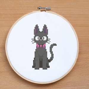 May include: A black cat with a pink bow tie is embroidered on a white canvas in a round wooden embroidery hoop.