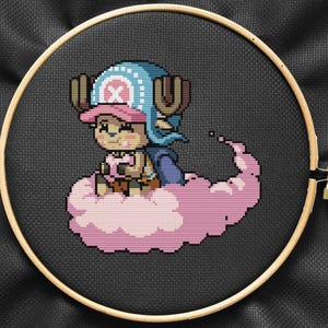 Puede incluir: Un bordado de punto de cruz con un personaje pixelado de la serie de anime One Piece, sentado en una nube rosa. El personaje lleva un sombrero azul, una gorra rosa y una capa azul. El bordado está en un aro de madera sobre un fondo de tela negra.
