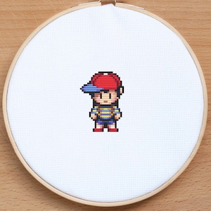 May include: Cross-stitch pattern of a pixelated character wearing a red and blue baseball cap, a yellow and blue striped shirt, and blue shorts.