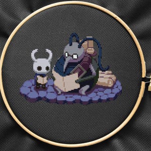 Peut inclure: Broderie au point de croix d'un personnage violet et gris du jeu vidéo Hollow Knight, tenant un livre. Un personnage blanc plus petit avec un masque noir se tient à côté du personnage plus grand.